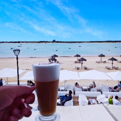 Vaso de cerveza con espuma, sostenido frente a la playa con sombrillas de paja