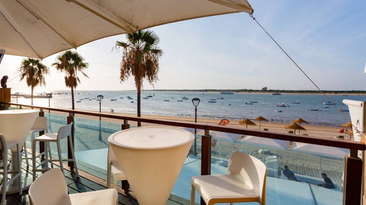 Terraza alta del bar con mesas blancas, palmeras y la playa de Bajo de Guía con barcas al fondo