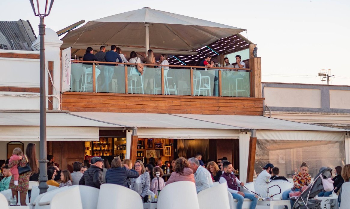 Terraza superior con barandilla de cristal llena de gente y, abajo, la terraza de sillones blancos